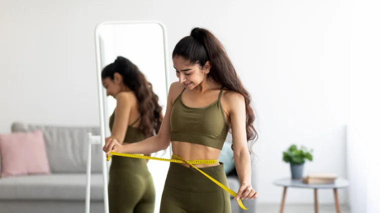 A person in front of a mirror measuring their waist size with a measuring tape.