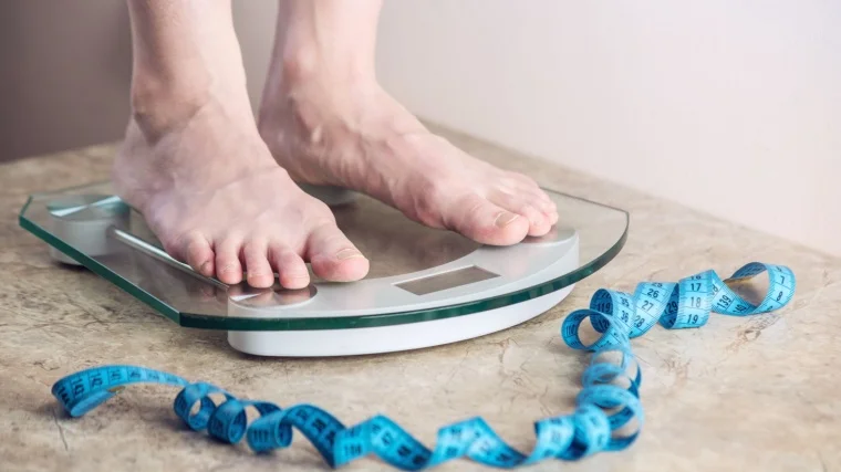 A person standing on an electronic weighing scale with a measuring tape on the floor.
