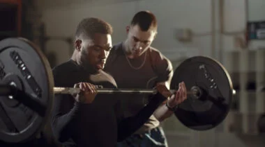 A person performs a barbell curl with their coach nearby.
