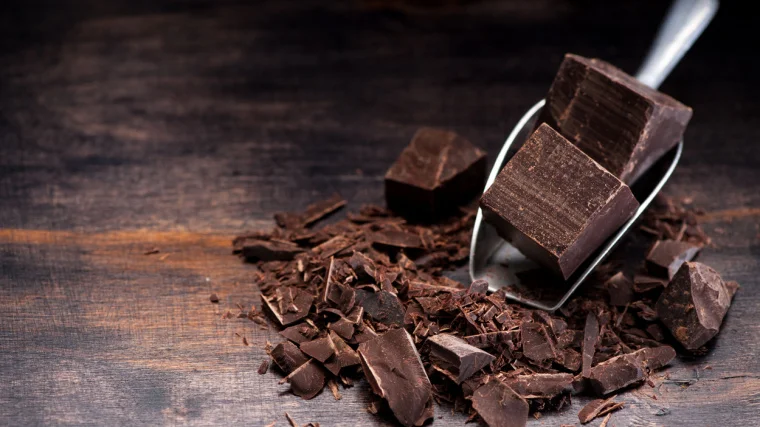Chunks of dark chocolate in a scoop and on a wooden surface.