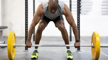 A person wearing a grey tank top prepares to lift a loaded barbell off the ground.