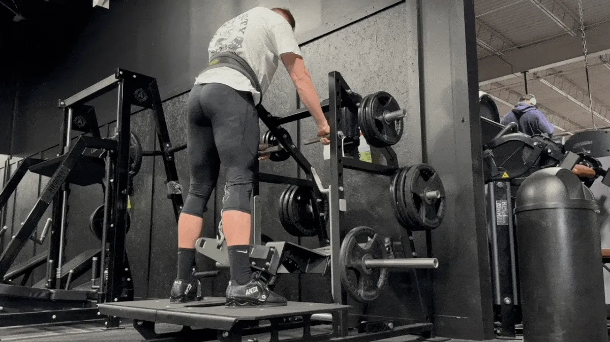 A person stands on a belt squat machine. A dip belt is secured around their waist, with a chain attached to one end of the belt and the other end to the machine. They hold the handles positioned in front of them, and then squat down before rising back up.