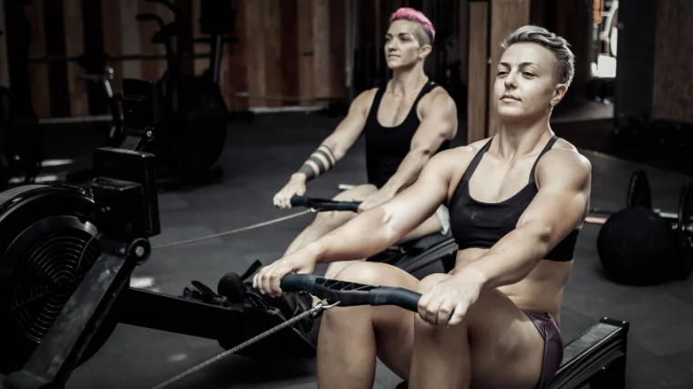 Two people with short hair wear a sports bra and tank top as they row on rowing machines.