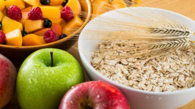 A bowl of fruit sits beside apples and a bowl of oats.