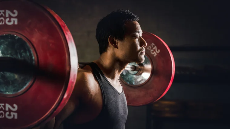 A person wears a black tank top and prepares to squat a barbell loaded with red 25kg plates.