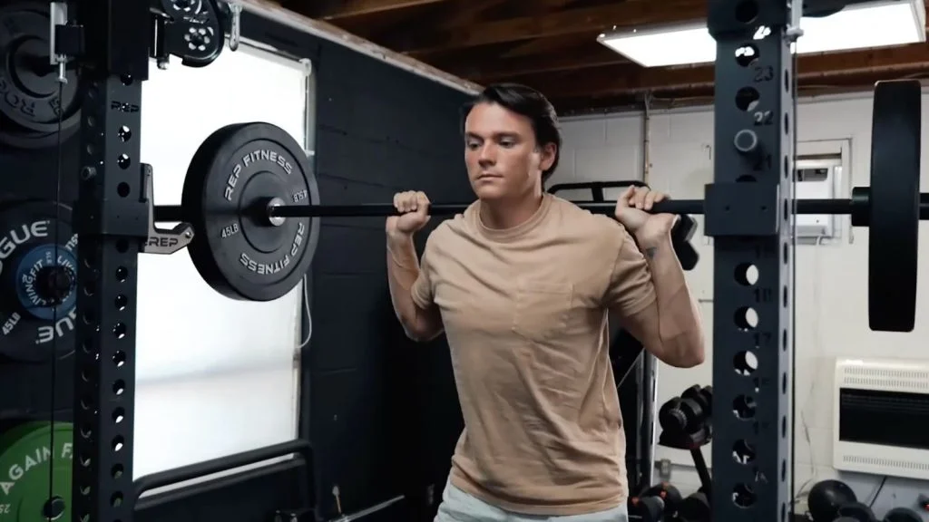 A person in a tan t-shirt squats with a loaded barbell draped across their back.