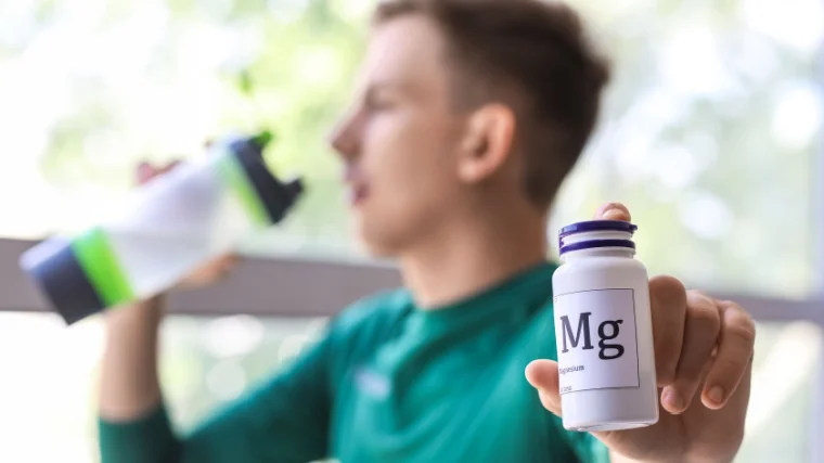 A person with a bottle of magnesium pills drinking water near a window.