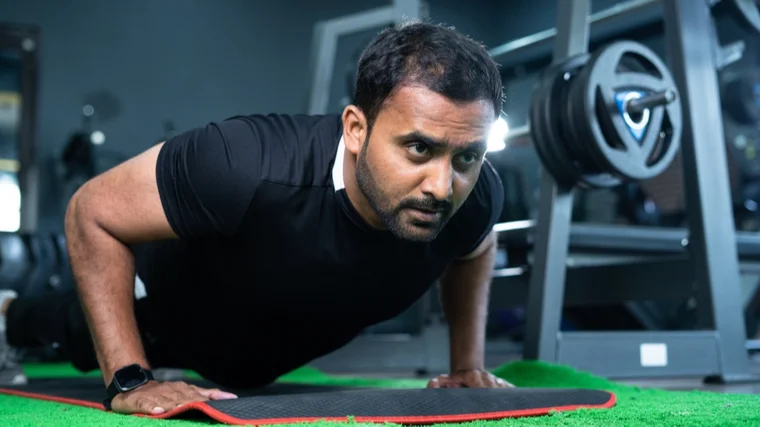 A person with facial hair wears a black t-shirt while performing a push-up in the gym.