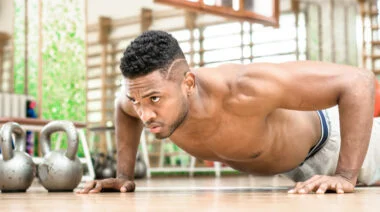 A shirtless person with short curly hair and some facial hair performs a push-up next to a pair of kettlebells.