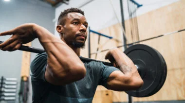 A person wears a blue-grey t-shirt and holds a loaded barbell in a front rack position.