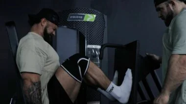 Bodybuilder Chris Bumstead, wearing a beige t-shirt, black shorts, and knee sleeves, performs seated leg presses as Iain Valliere spots him