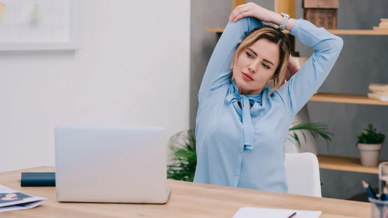 A woman seated in home office in front of computer stretching shoulder above head.