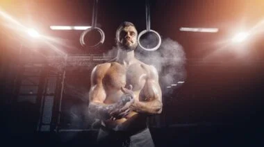 Shirtless person with hands covered in chalk standing in front of a set of rings.