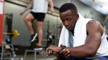 An athlete with a white towel draped around their shoulders sits in the gym while checking their watch.