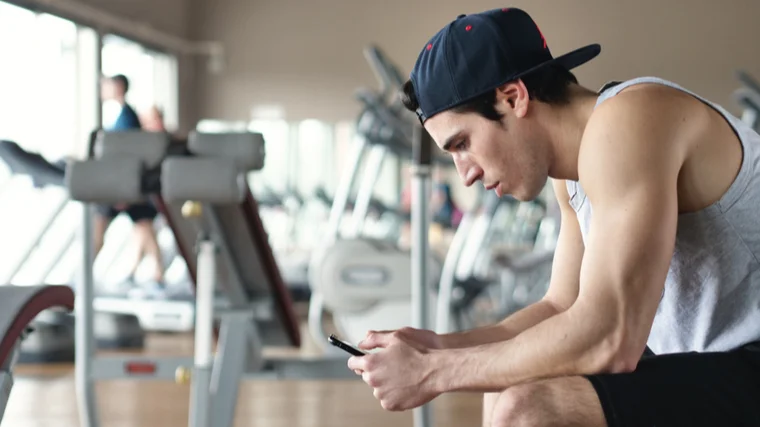 A person wears a backwards baseball cap and sits on a gym bench while looking at their smartphone.