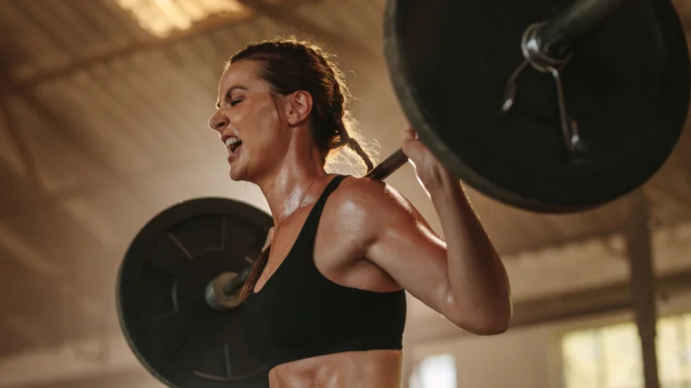 A person wearing a tank top yells in exertion as they hold a loaded barbell on their back.