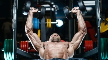 A muscular person performing the dumbbell bench press exercise in the gym.