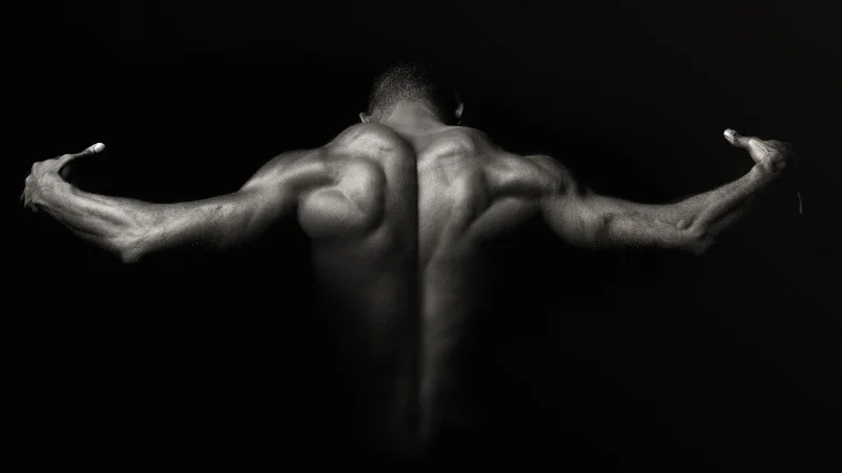 An athlete in a dark room flexing back and shoulder muscles with light accenting muscle form and build.