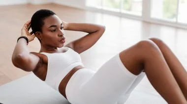A person wearing a white sports bra and workout shorts performs a crunch in a yoga studio.