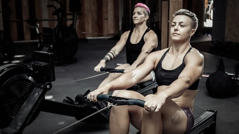 Two people with short haircuts use rowing machines in the gym.