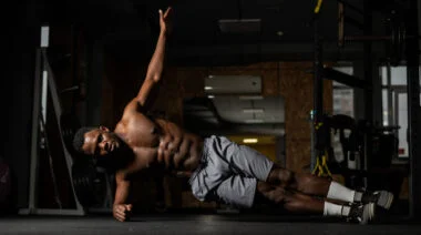 A shirtless person performs a side plank with their upper arm extended in the gym.