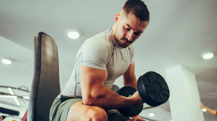 A bodybuilder sitting on a weight bench curling their biceps with a dumbbell.