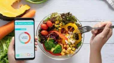 Fresh fruits and vegetables in a bowl and a hand holding a smartphone showing a food calorie tracker.