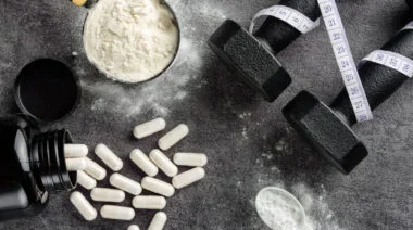 Protein and BCAA powder in scoops next to a set of dumbbells and measuring tape.