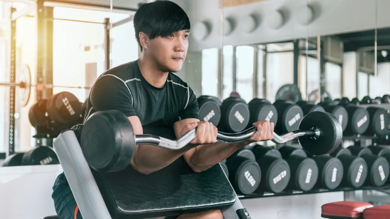 A person wears a black t-shirt with silver piping as they perform a preacher curl with a curl bar.