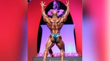 Bodybuilder Phil Heath stands on the Mr. Olympia stage, his back facing the audience and his arms extended over his head. he's wearing sparkly blue posing trunks.