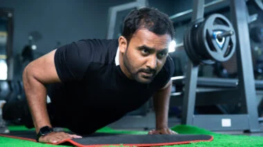 A person with facial hair wears a black t-shirt while performing a push-up in the gym.