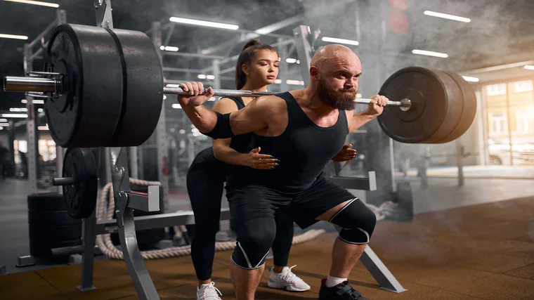 A person wears a tank top and knee sleeves while squatting heavy and receiving a spot from someone behind him.