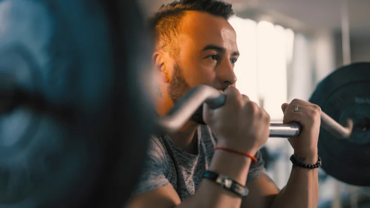 A close-up shows a person with facial hair wearing a few bracelets while performing a preacher curl with a loaded curl bar.