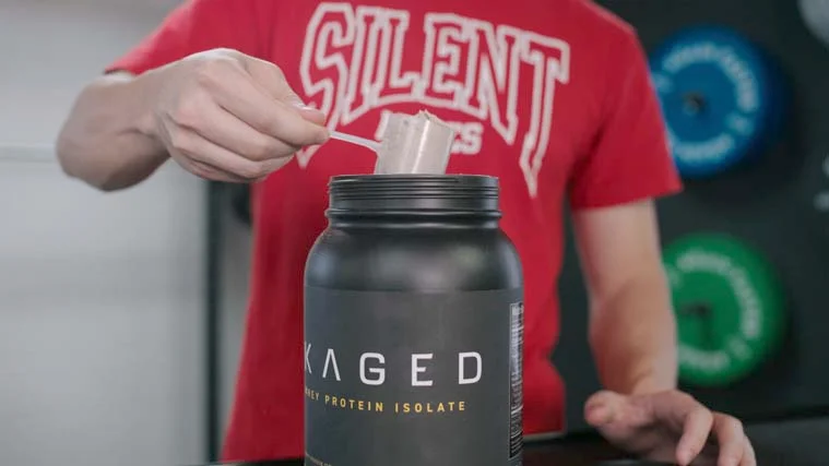 A person in a red shirt holding up one Scoop of Kaged Whey Protein Isolate.