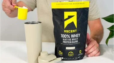a person holding a scoop of Ascent Whey Protein over a shaker cup