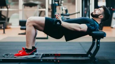 A man laying his upper back against a weight bench and performing a hip thrust with a barbell across his lap.