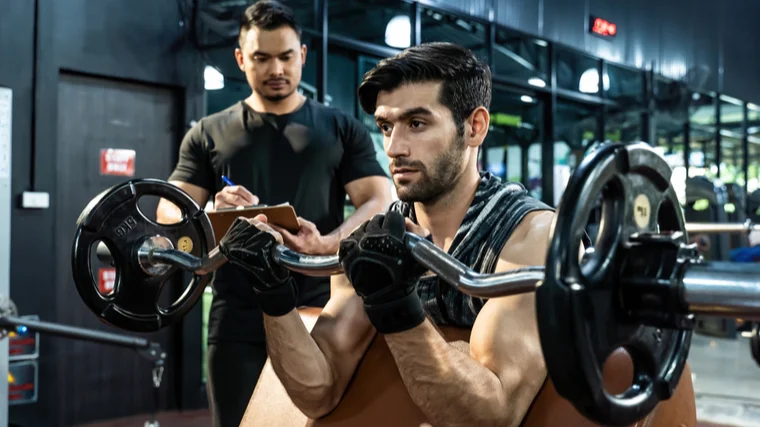 A person performs an EZ-bar curl with their coach looking on.