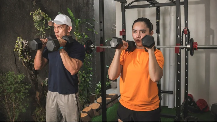 Two people wearing t-shirts and workout shorts hold dumbbells ready to press overhead.