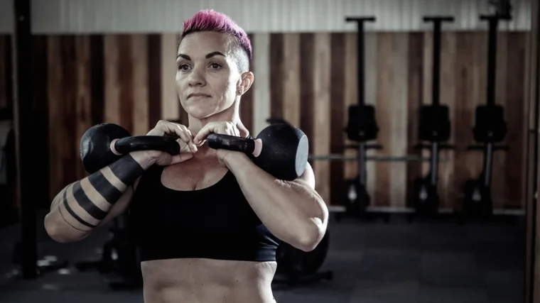 A person with short pink hair and forearm tattoos wears a sports bra while holding two kettlebells in front rack position.