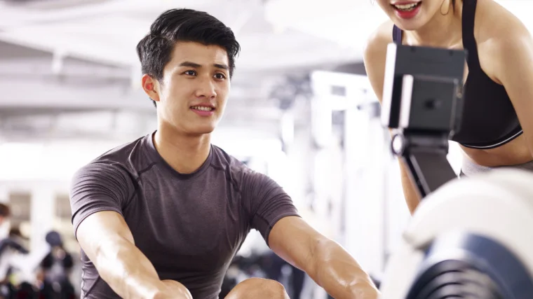 A person works out on a rowing machine while a coach encourages them.