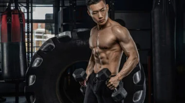 A muscular shirtless person looks at themself in the mirror from the side while performing hammer curls.